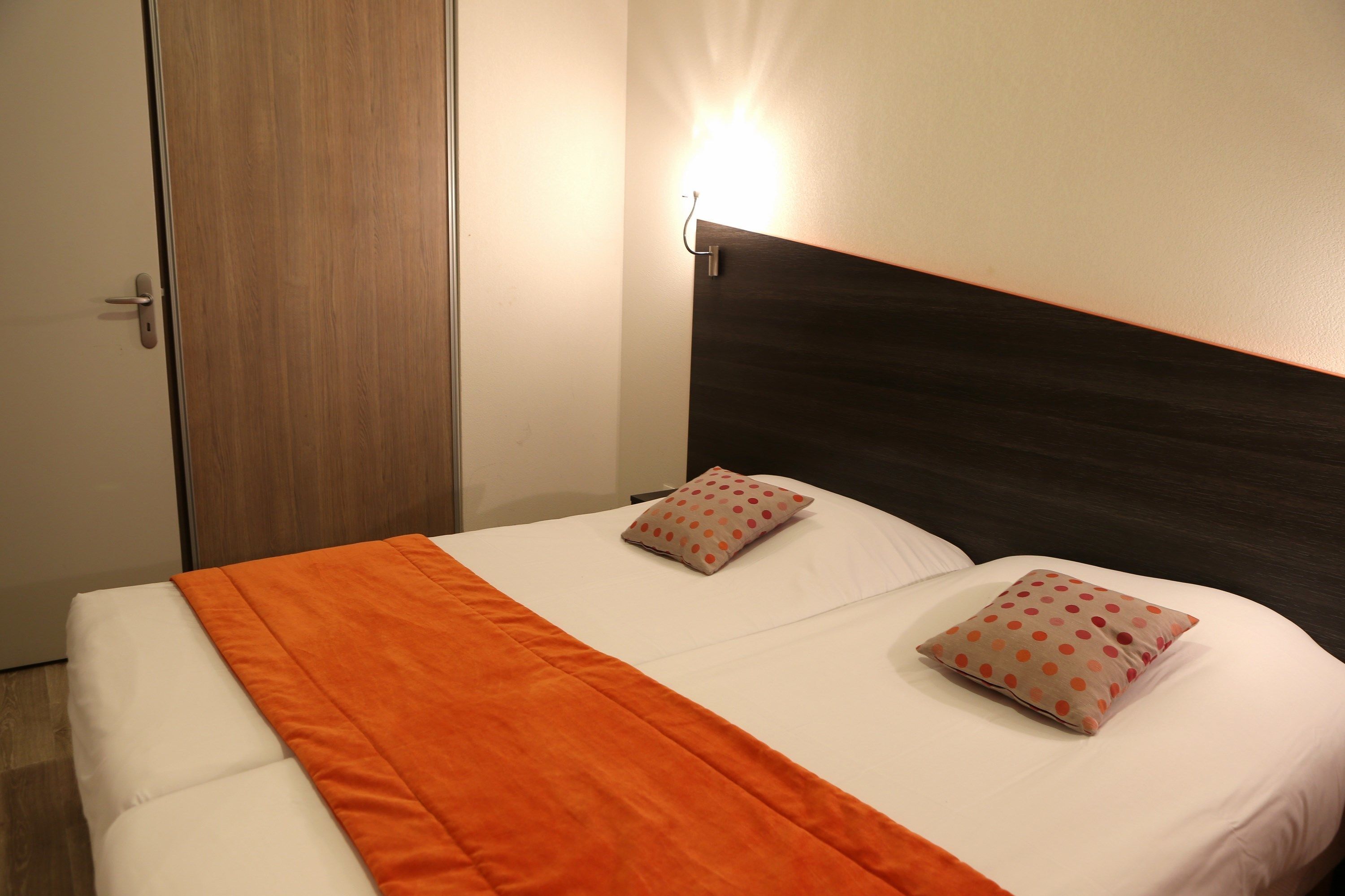 Una cama bien hecha con un edredón naranja, dos almohadas con lunares y una lámpara de pared.