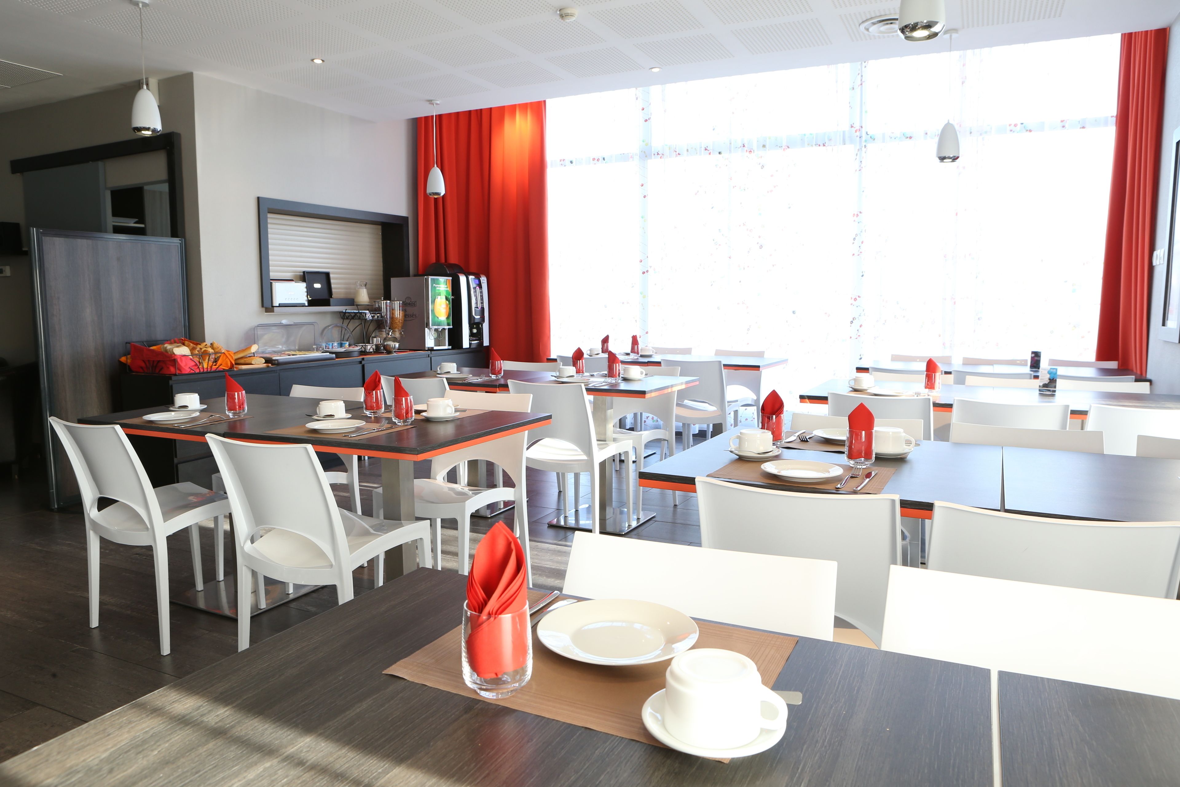 Un comedor moderno con varias mesas y sillas, una zona de cocina con electrodomésticos, y ventanas grandes con cortinas rojas.