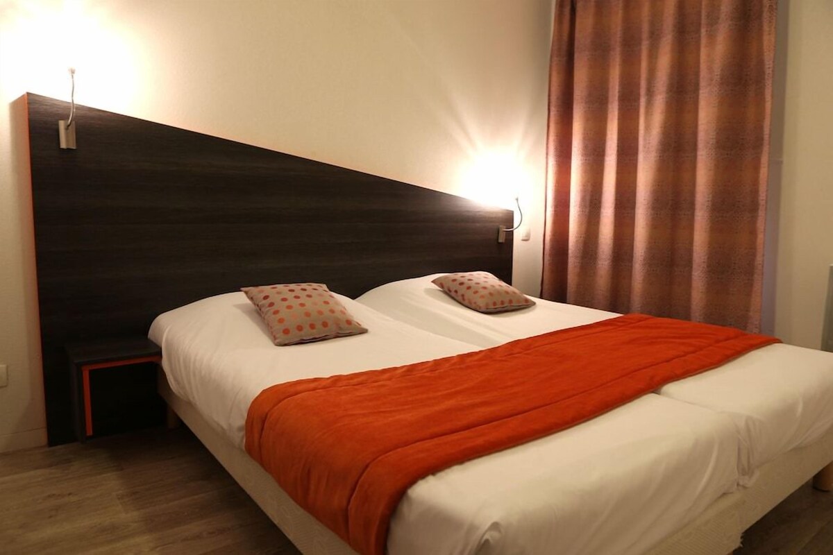 A double bed with an orange blanket, two pillows, a nightstand, and a wall-mounted light.