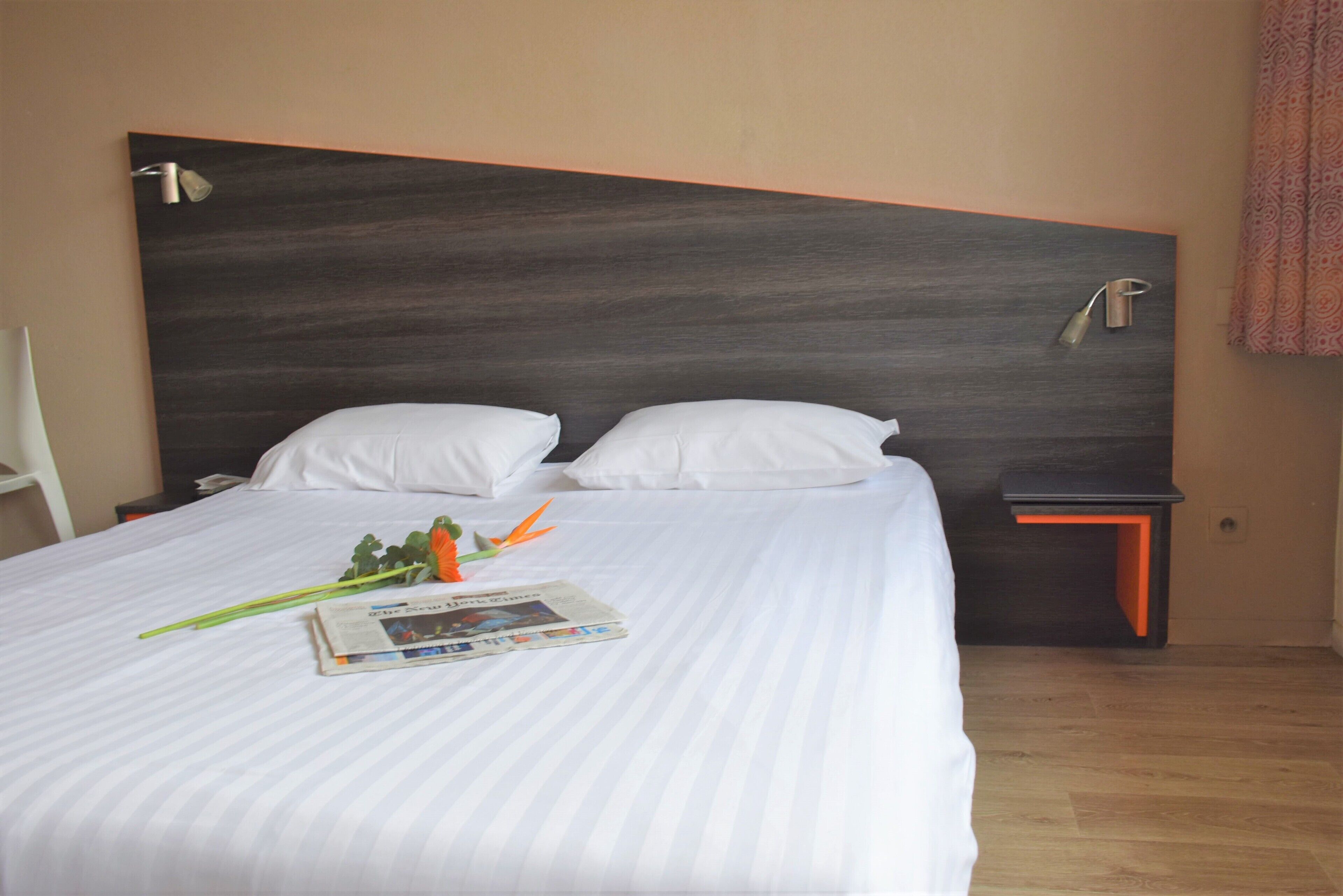 Habitación de hotel con una cama grande, dos almohadas, una mesita de noche, una lámpara y un periódico sobre la cama.