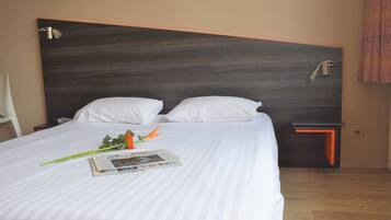 Habitación de hotel con una cama grande, dos almohadas, una mesita de noche, una lámpara y un periódico sobre la cama.