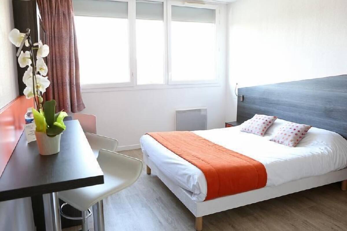 Quarto de hotel com uma cama, escrivaninha, cadeira, planta em vaso e janela com cortinas.