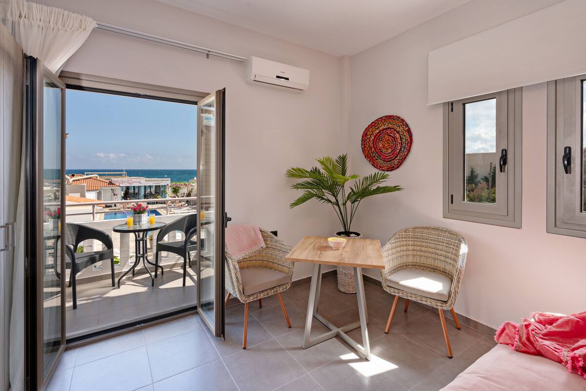 A balcony with a view of the sea and buildings, wicker chairs, a small table, and a potted plant.