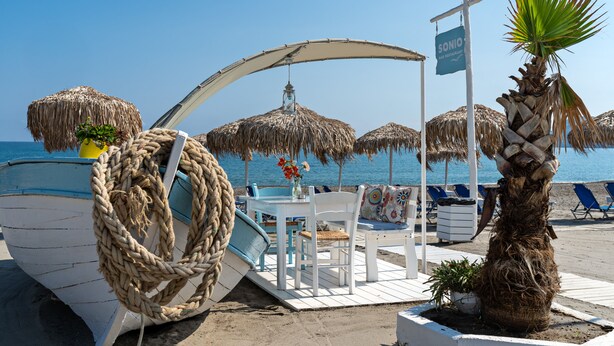 Un restaurante frente al mar con un bote, sombrillas de paja y sillas reclinables.