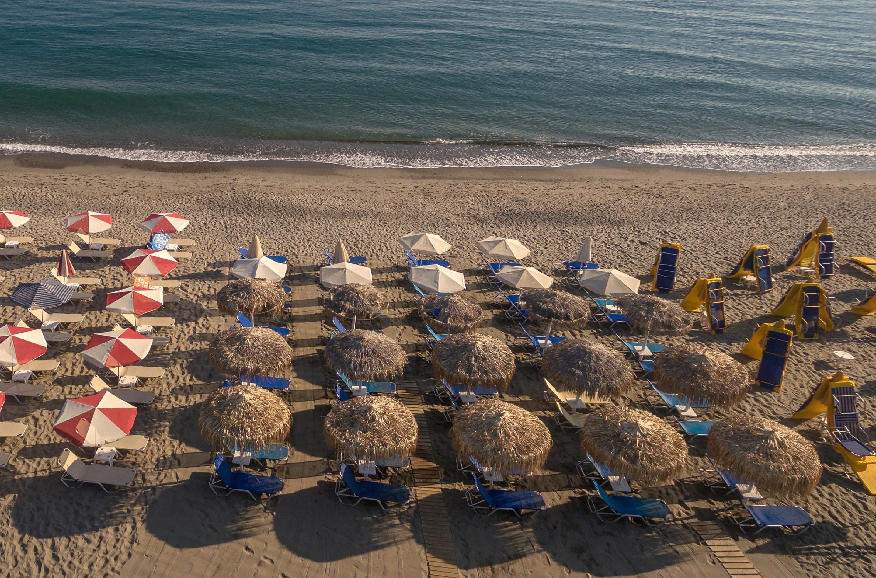 A beach with rows of sun loungers and umbrellas, some with thatched roofs, and a calm sea in the background.