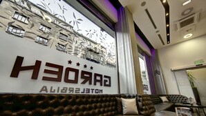 Een moderne hotel lobby met een leren bank, een groot bord met de tekst 'GARDO HOTEL' en een decoratief raam met bladmotieven.