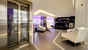 Een moderne hotellobby met een receptiebalie, een zithoek en een glazen lift.