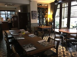 A restaurant with a long wooden table, multiple chairs, and a patterned floor. There is a counter with a person at it, and a window with curtains.