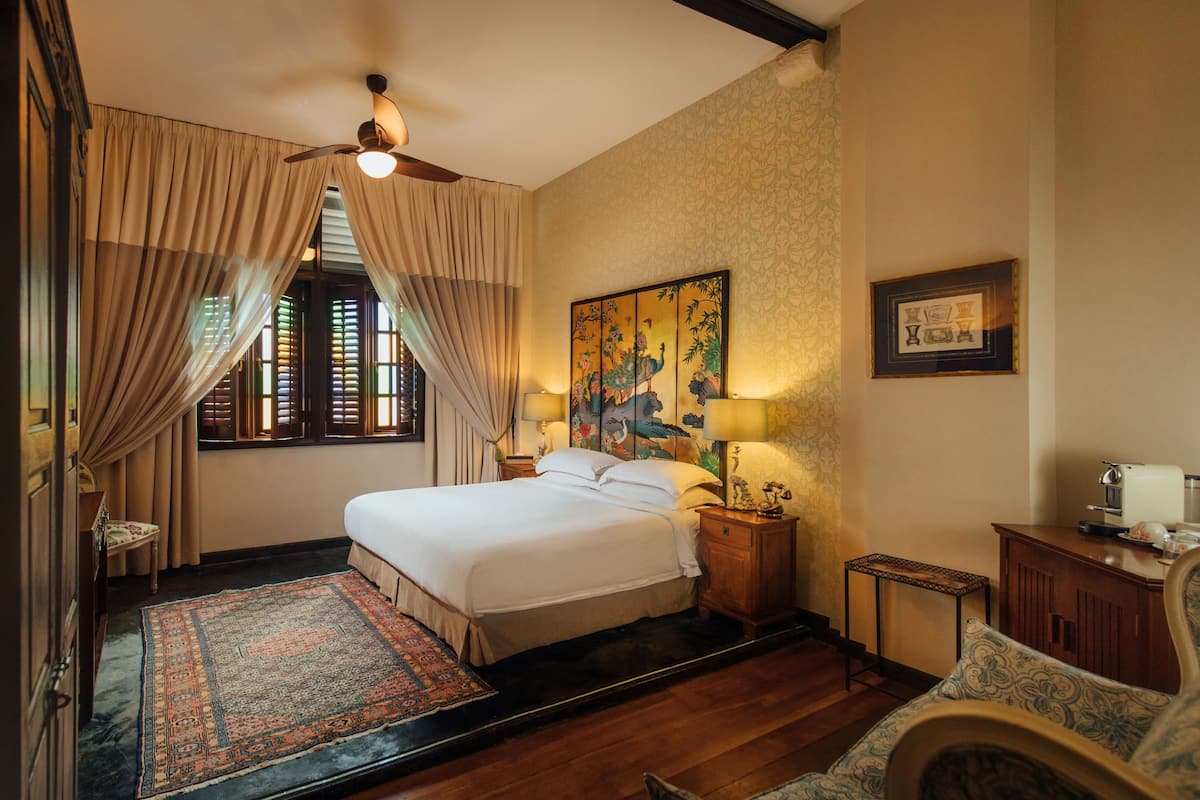 A hotel room with a large bed, a wooden floor, a traditional rug, a desk with a lamp, and a painting on the wall.