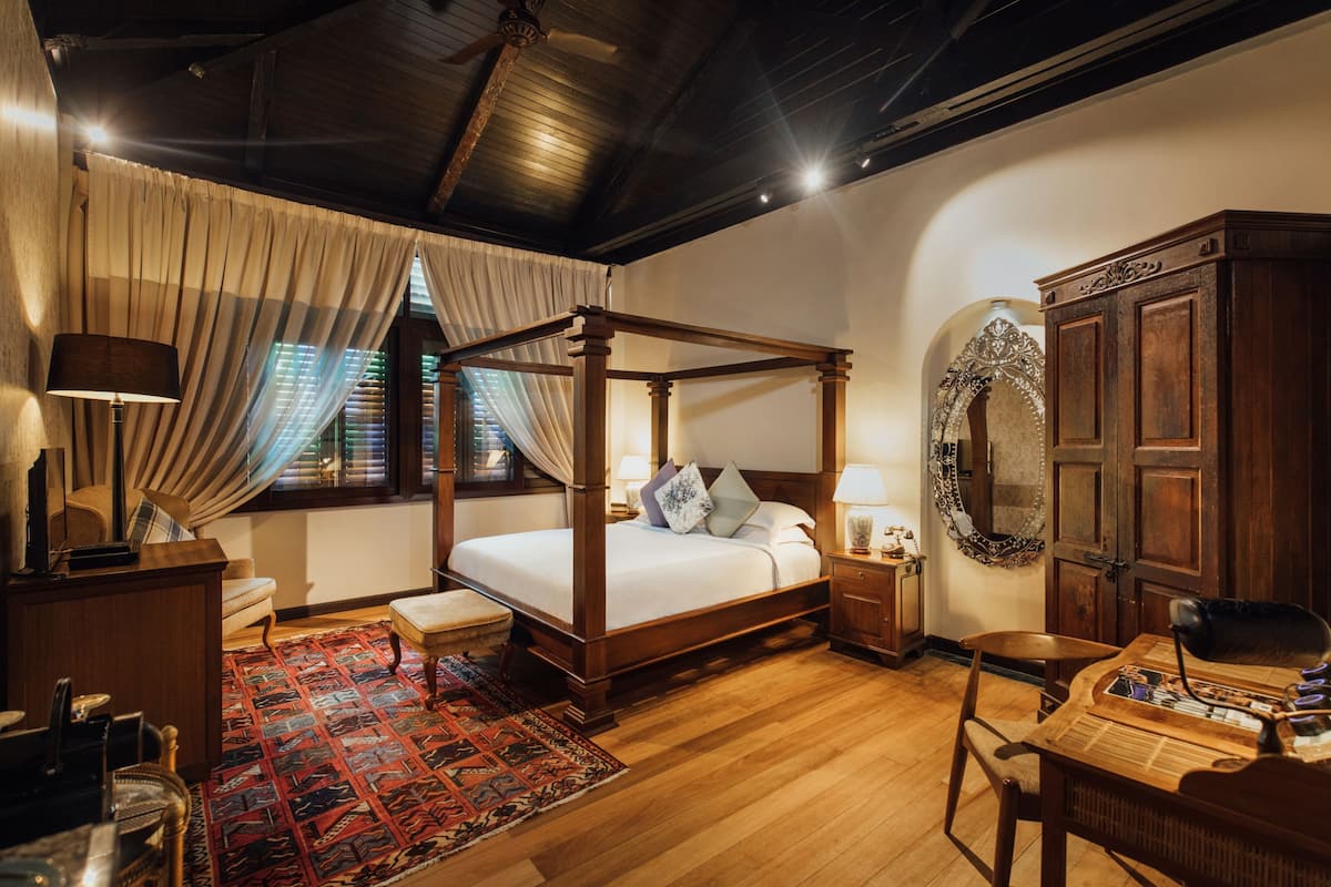A four-poster bed with a wooden frame, a desk with a chair, a desk lamp, and a patterned rug.