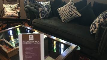A sign advising social distancing practices in a room with a glass table, a sofa with patterned cushions, and a wooden chair.