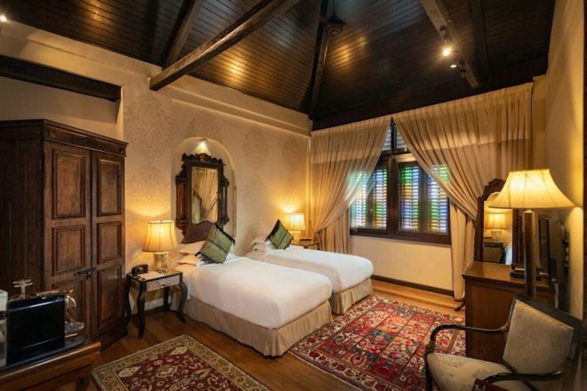 Une chambre d’hôtel avec deux lits, un plafond en bois, une grande fenêtre avec des rideaux et un tapis traditionnel.