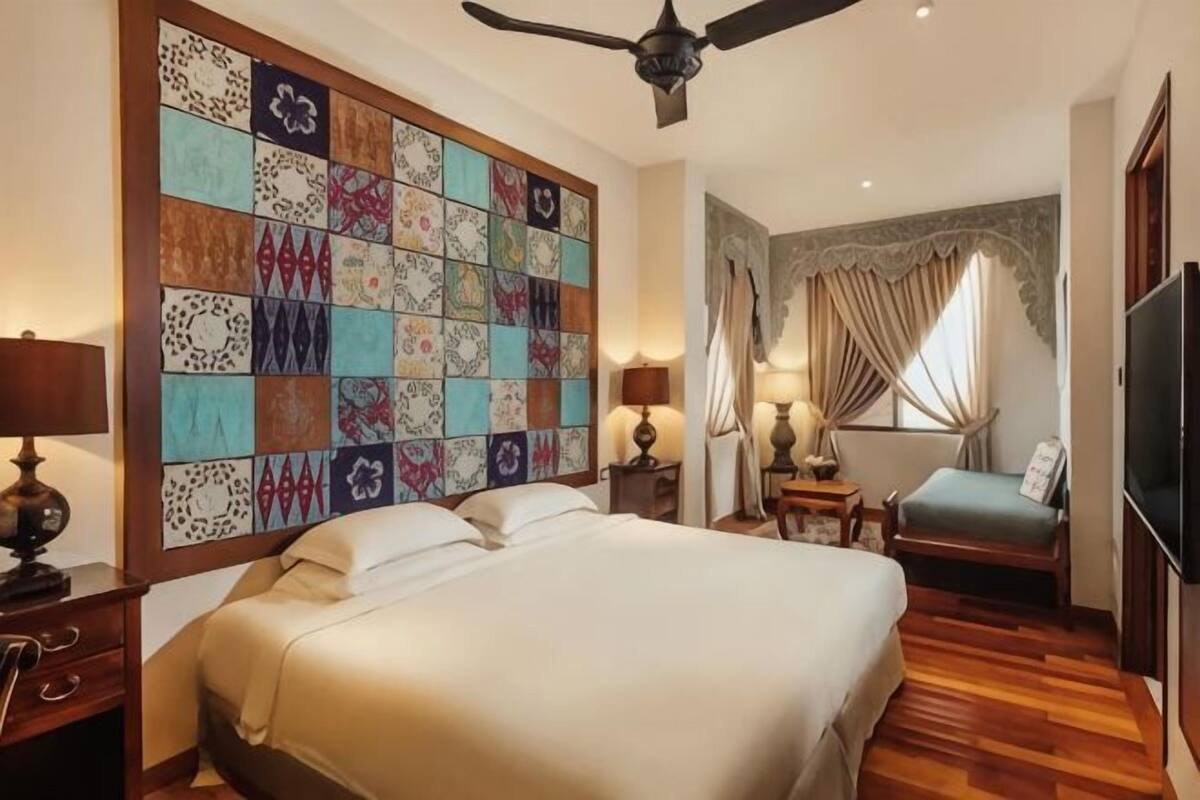 A hotel room with a large bed, a ceiling fan, a television, a desk with a lamp, and a decorative wall panel.
