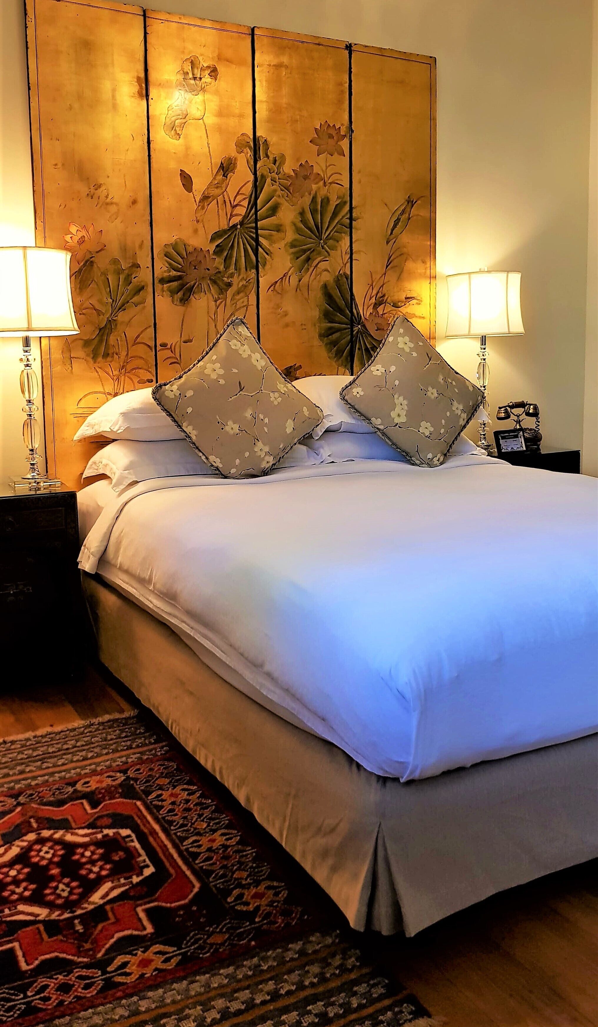 Un lit bien fait, avec du linge de lit blanc et des coussins à motifs floraux, flanqué de deux lampes de chevet.