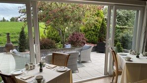 Un coin repas avec une table dressée pour quatre personnes, donnant sur un jardin où poussent différentes espèces de plantes et un grand arbre aux fleurs roses.