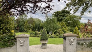 Un portail en pierre, flanqué de deux statues de lions en pierre, mène à un jardin bien entretenu, agrémenté d’un parterre central et d’un chemin pavé.