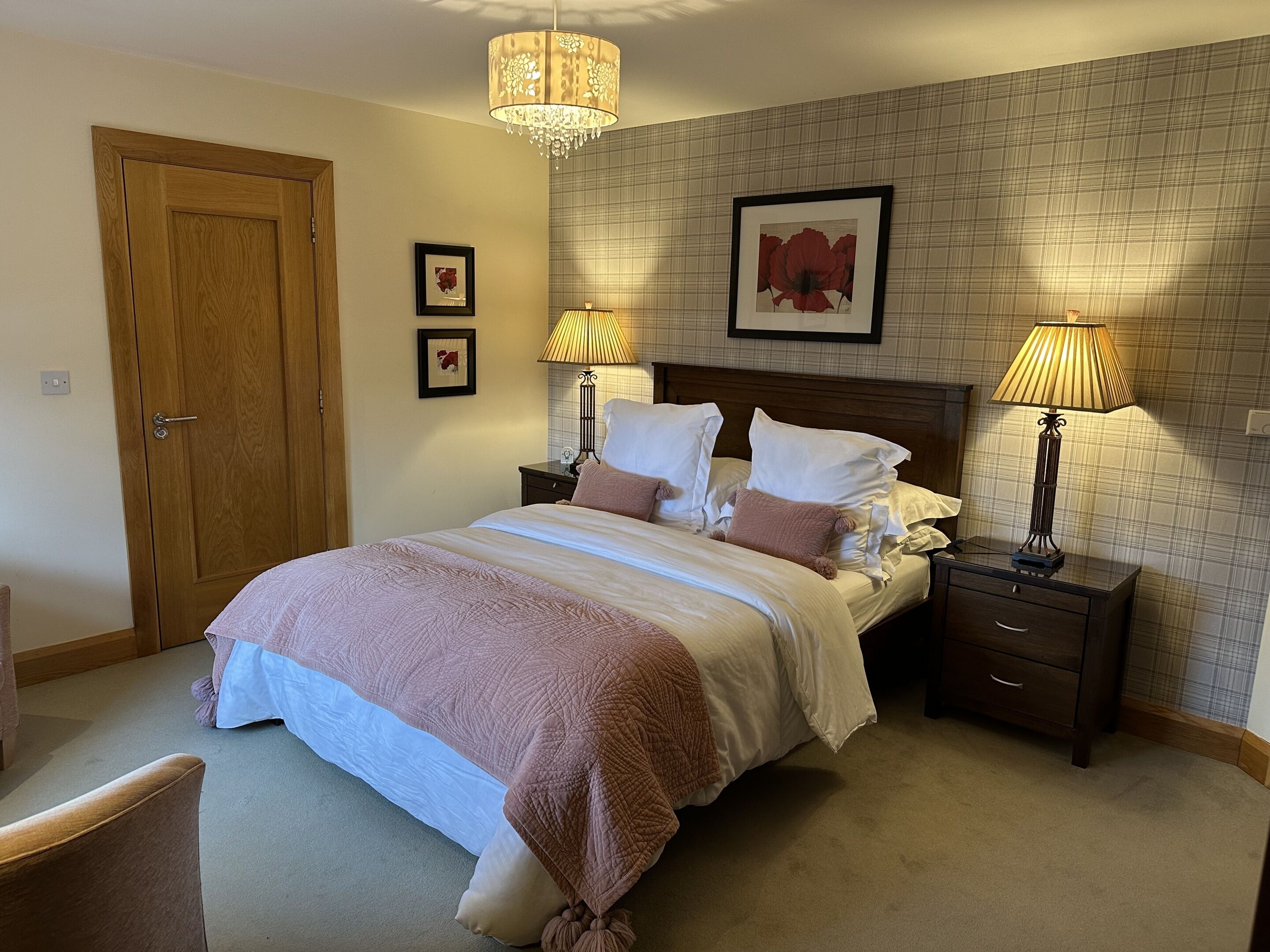 A neatly made bed with a white and pink bedspread, two bedside lamps, a wooden headboard, and a plaid-patterned wall.