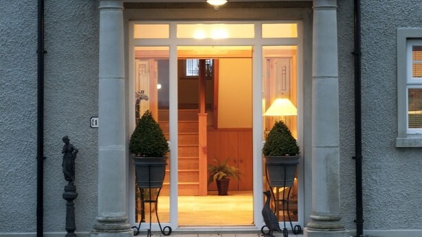 Une entrée d’immeuble avec un luminaire au-dessus de la porte, deux plantes en pot de chaque côté et une statue à gauche.