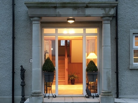 A building entrance with a light fixture above the door, two potted plants on either side, and a statue on the left.