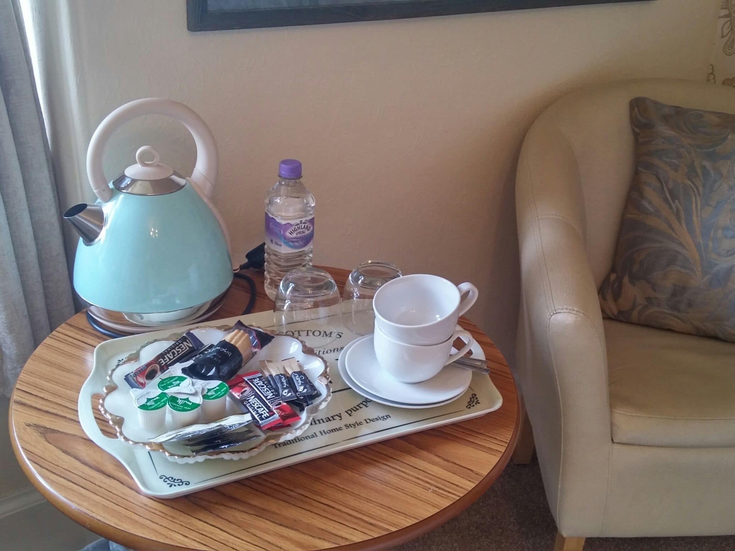 A tray with tea bags, a teapot, a bottle of water, and a cup on a round table in a room with a sofa and a pillow.