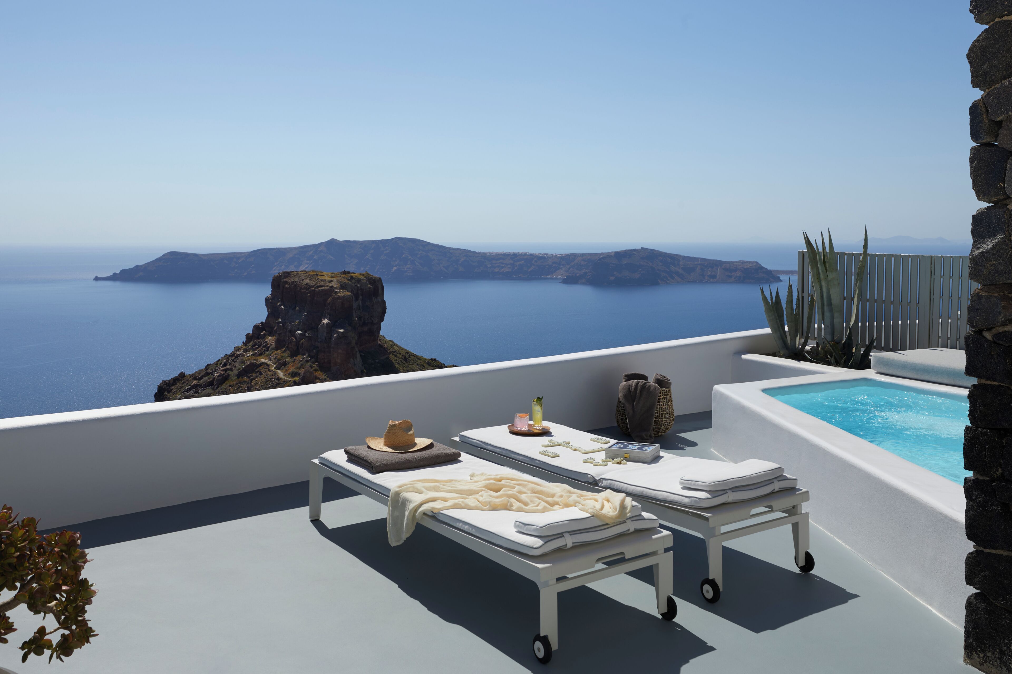 A sunlit terrace with two sunbeds, a small table with a hat and candles, and a stone wall.