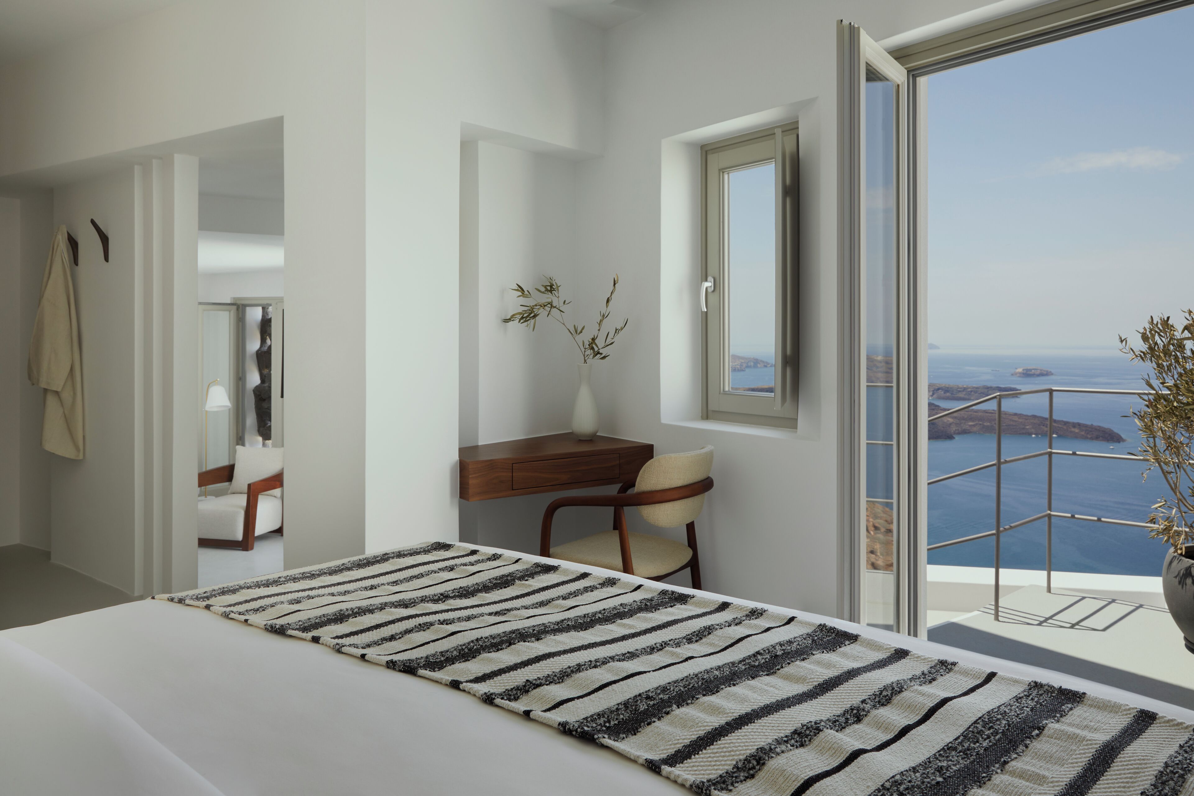 A modern bedroom with a large bed, a desk, and a chair. There is a view of the sea through a balcony.