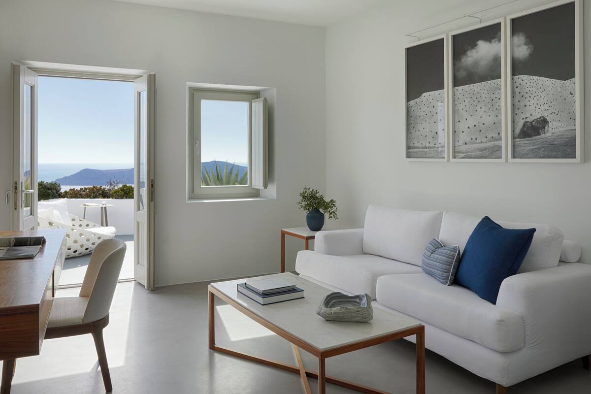 A modern living room with a white sofa, a coffee table, and a dining area with a wooden table and chairs. There are two windows with views of the outdoors.