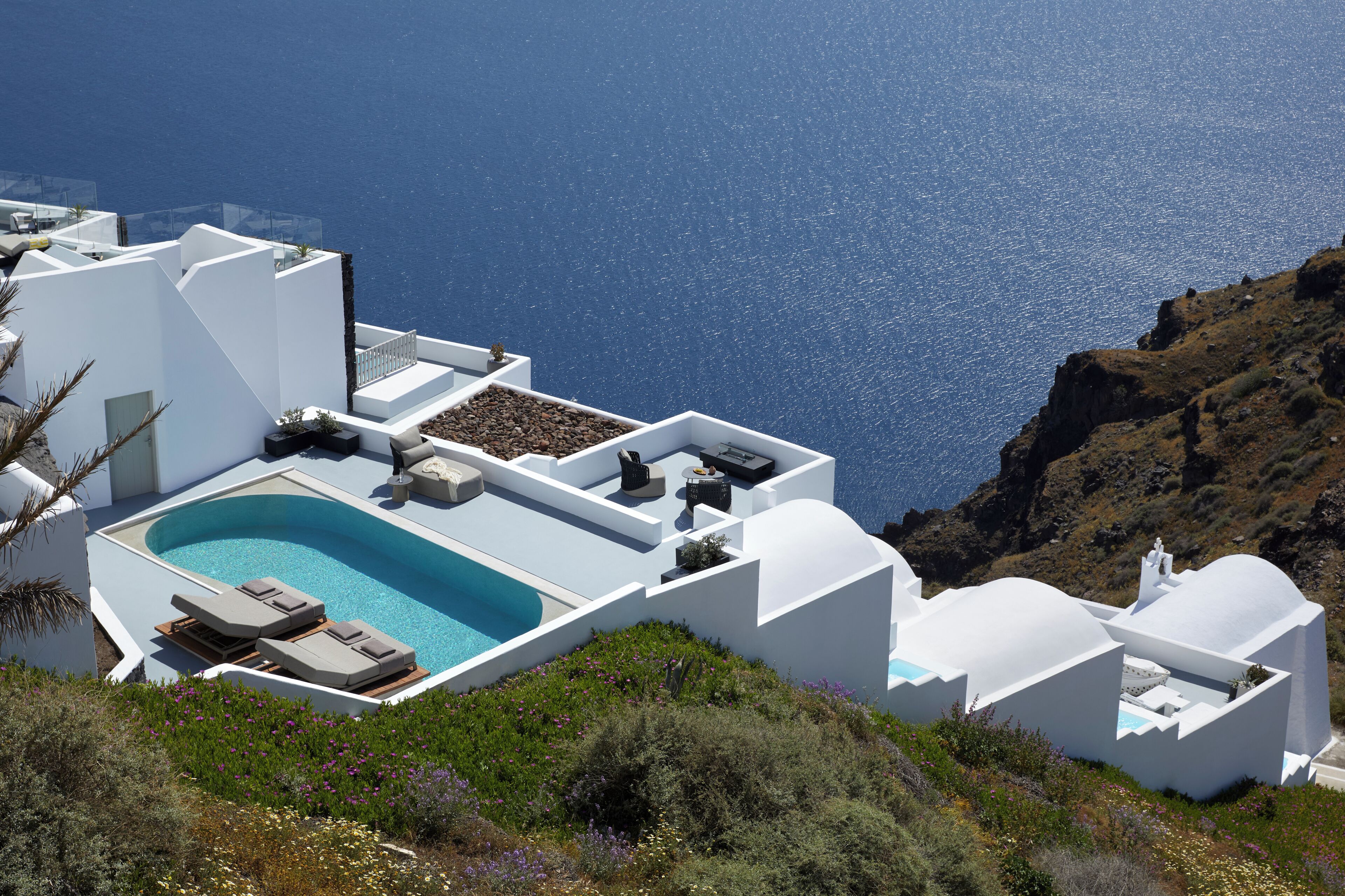 Een modern wit gebouw met een zwembad, lounge stoelen en uitzicht op zee.