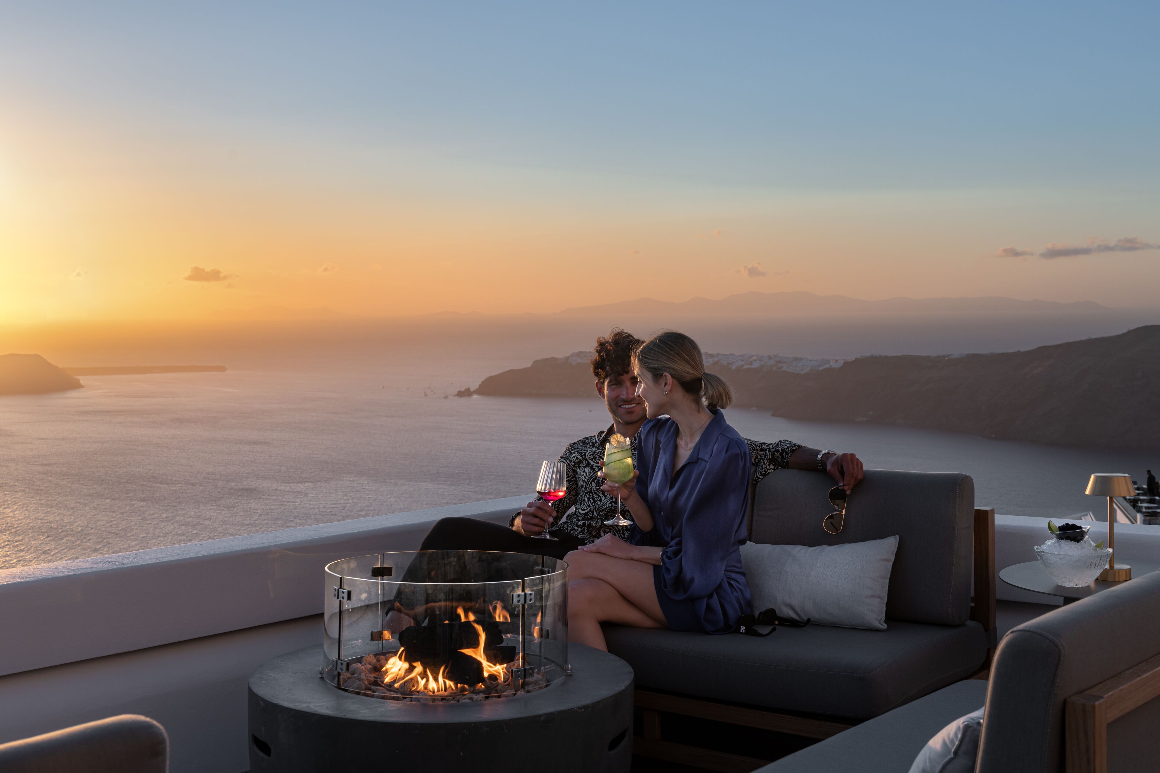 A couple sitting by a fire pit with a view of the sea and mountains during sunset.
