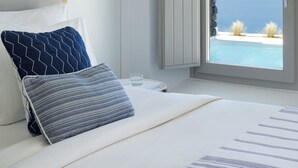 A bedroom with a bed, pillows, a bedside table, a hanging light, and a view of the sea through an open door. - Grace Hotel, Auberge Collection (Santorini)