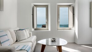 A modern living room with a white sofa, a round table, and two windows offering a view of the sea. - Grace Hotel, Auberge Collection (Santorini)