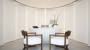 A reception area with a reception desk, two wooden chairs, a white vase with flowers, and a computer monitor.