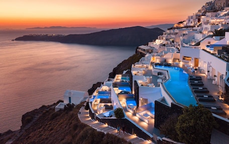 A resort with a pool, white buildings, and a view of the sea at sunset.