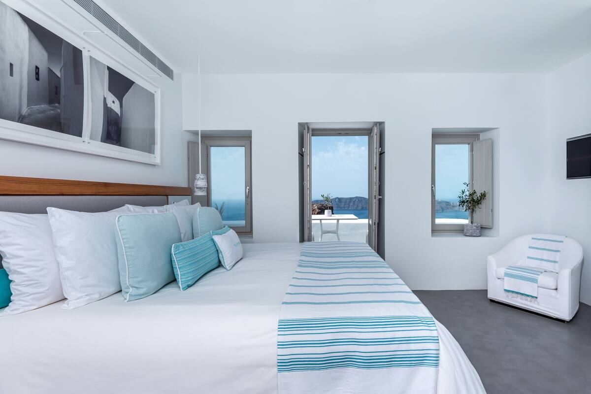 A modern hotel room with a large bed, two windows, a view of the sea, a chair, and a small table with a plant.