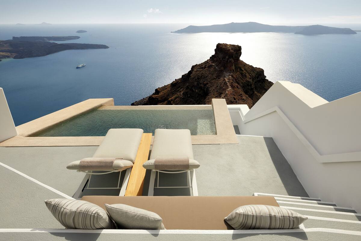A rooftop infinity pool with two lounge chairs and a wooden deck, overlooking a scenic view of the sea and islands.