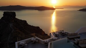 A sunset view from a rooftop terrace with tables and chairs, overlooking a calm sea and rocky coastline. - Grace Hotel, Auberge Collection (Santorini)