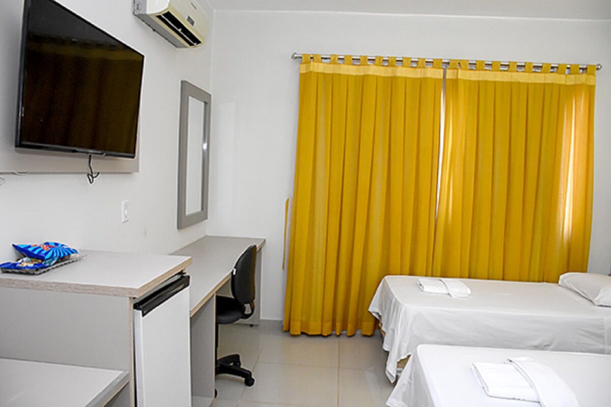 Een hotelkamer met een bureau, twee bedden, een televisie en een geel gordijn.
