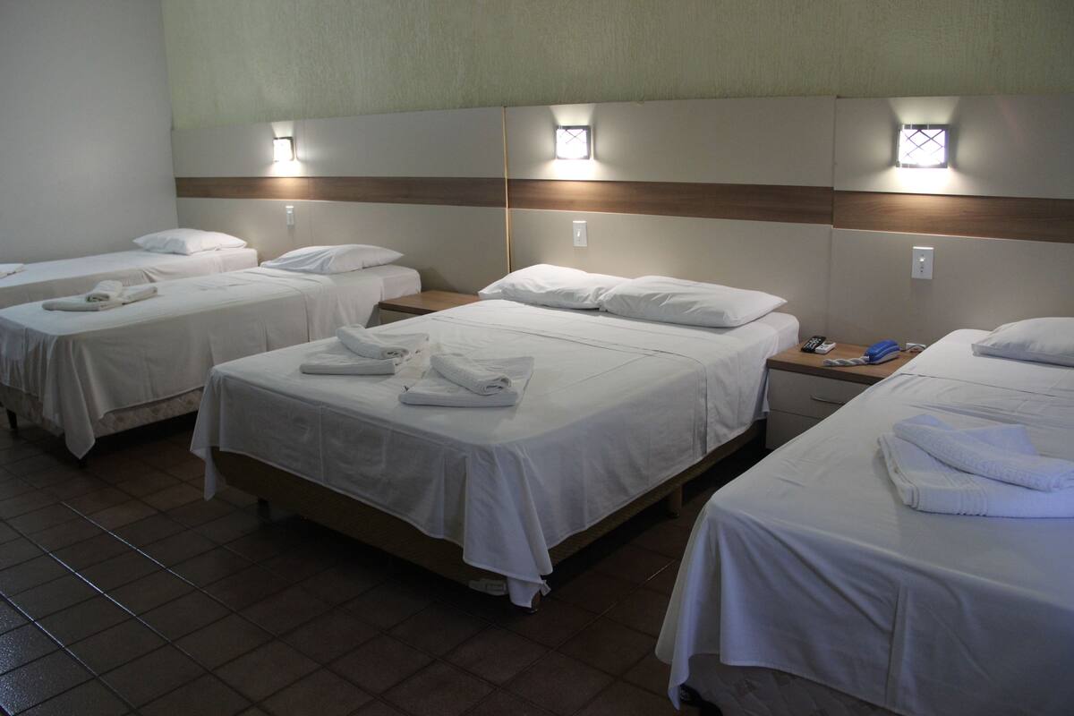 Habitación de hotel con tres camas individuales, cada una con cubrecama y almohadas blancas, y una mesita de noche con lámpara.