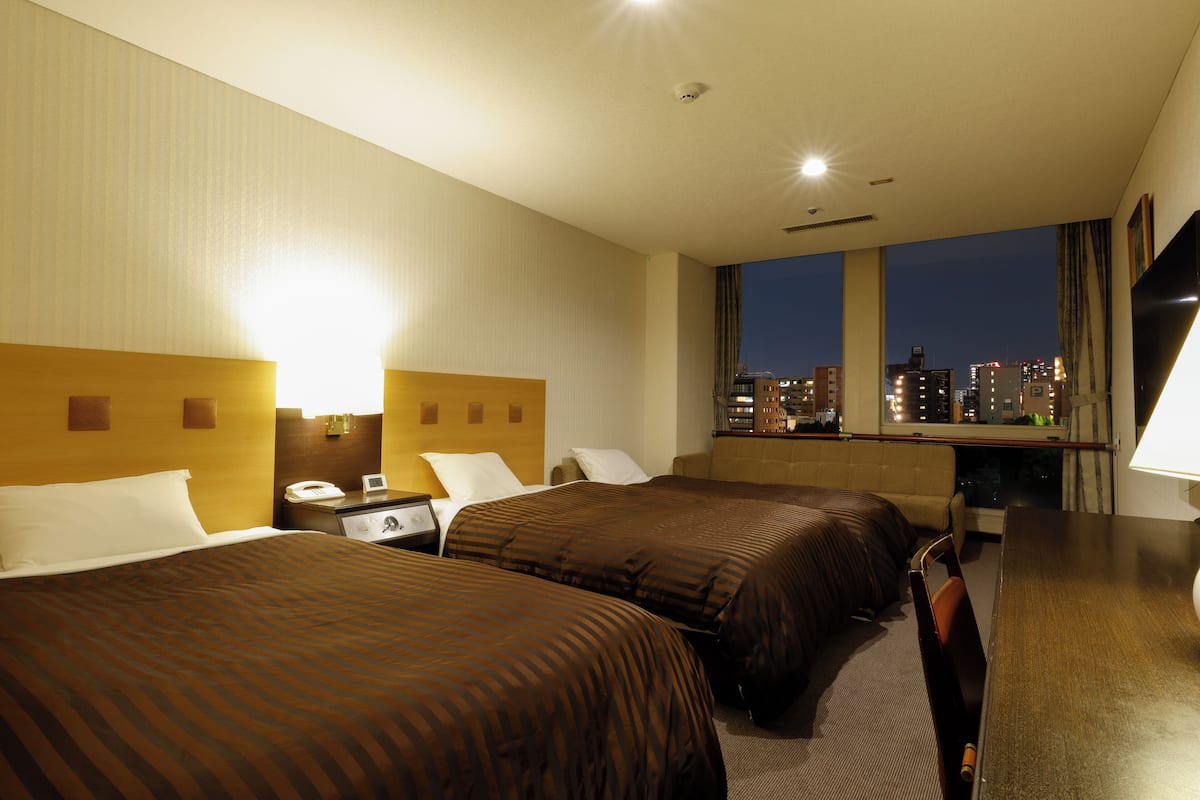 A hotel room with two beds, a desk, a chair, a TV, and a window with a city view.