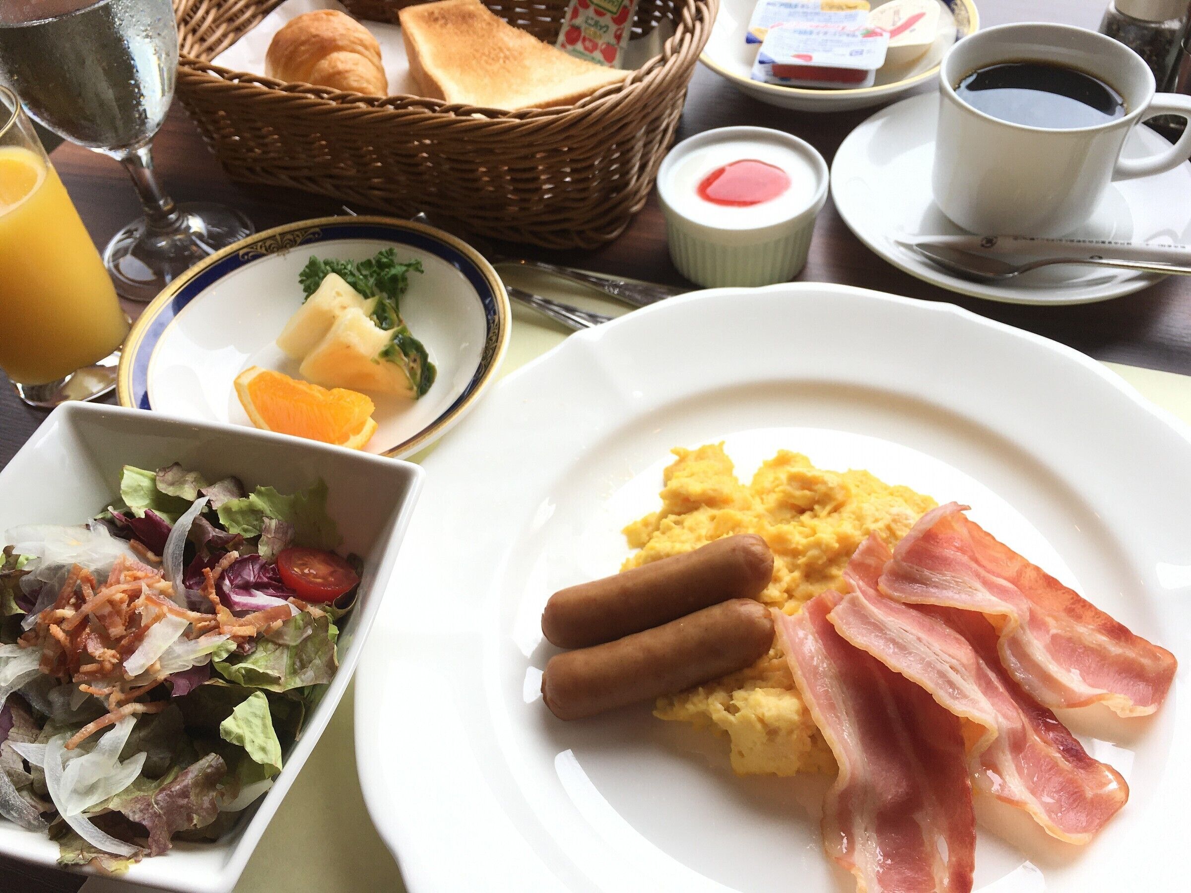 Een ontbijttafel met roerei, spek, worstjes, toast, een salade en een kop koffie.
