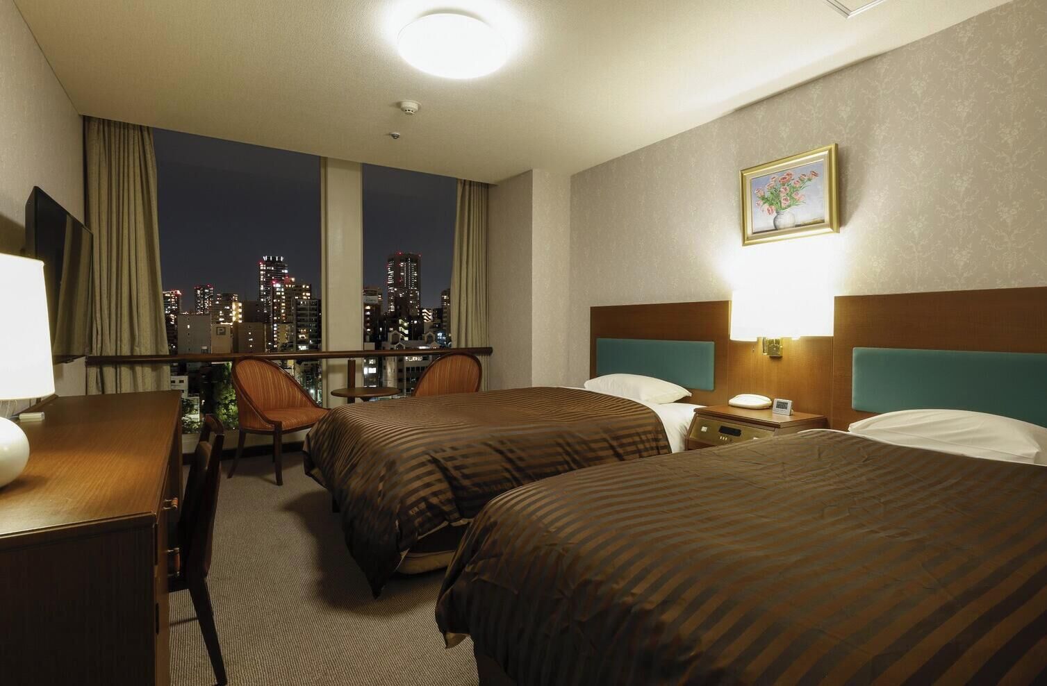 Une chambre d’hôtel avec deux lits, un bureau, une chaise, une fenêtre donnant sur la ville et un tableau au mur.