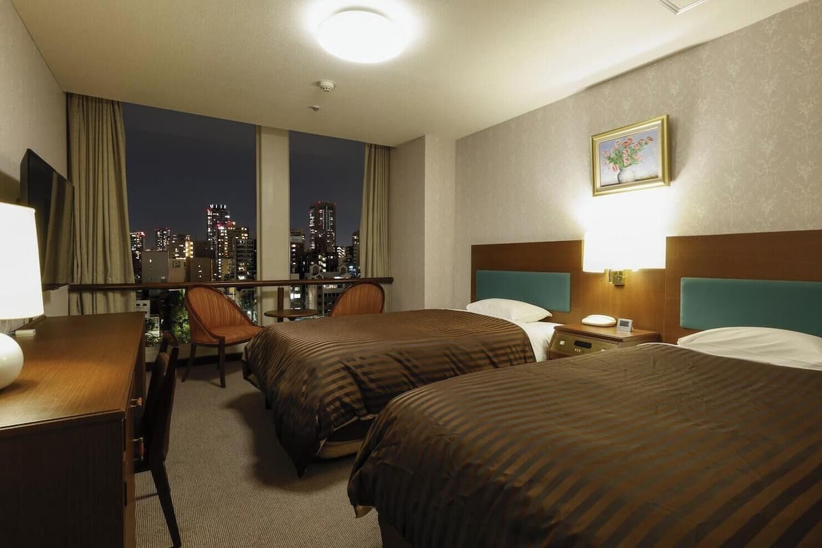 A hotel room with two beds, a desk, a chair, a window with a city view, and a painting on the wall.