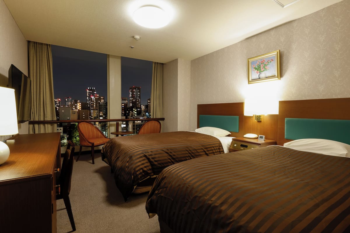 A hotel room with two beds, a desk, a chair, and a view of the city skyline.