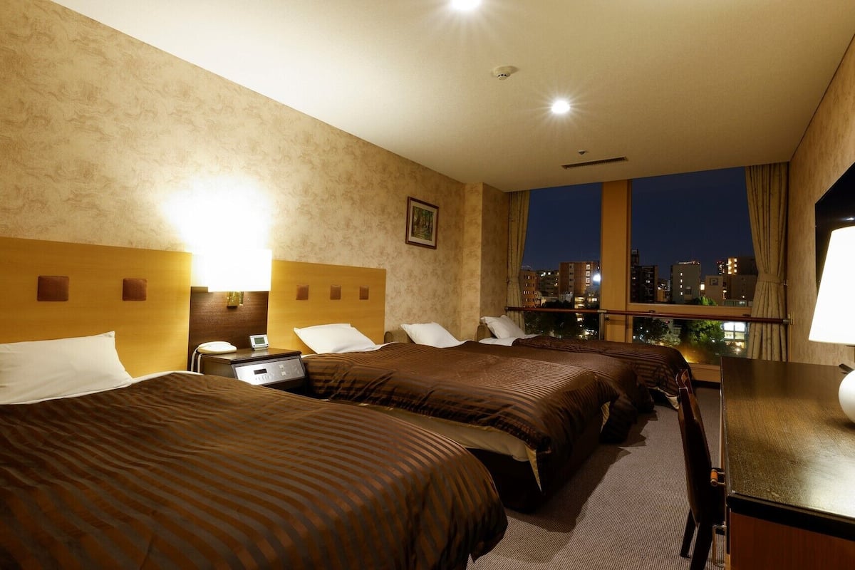A hotel room with three beds, a desk, a chair, a window with a city view, and a ceiling light.