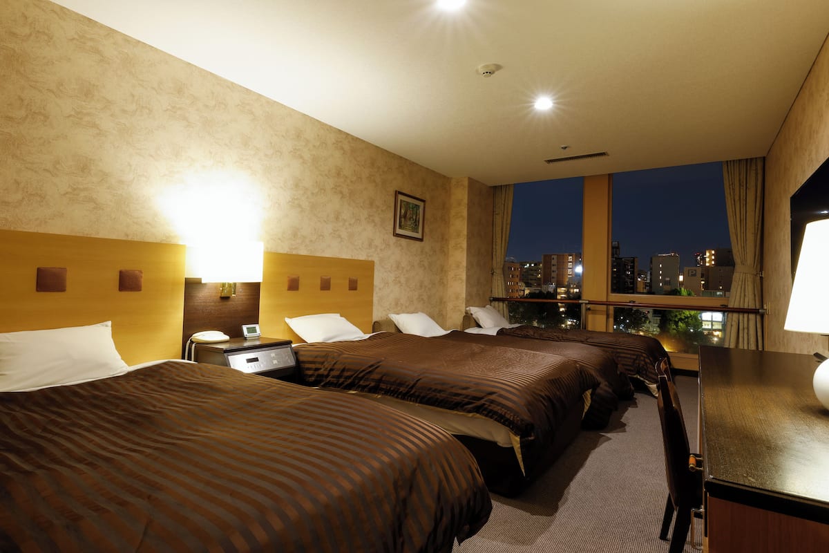 A hotel room with three beds, a desk, and a view of the city at night.