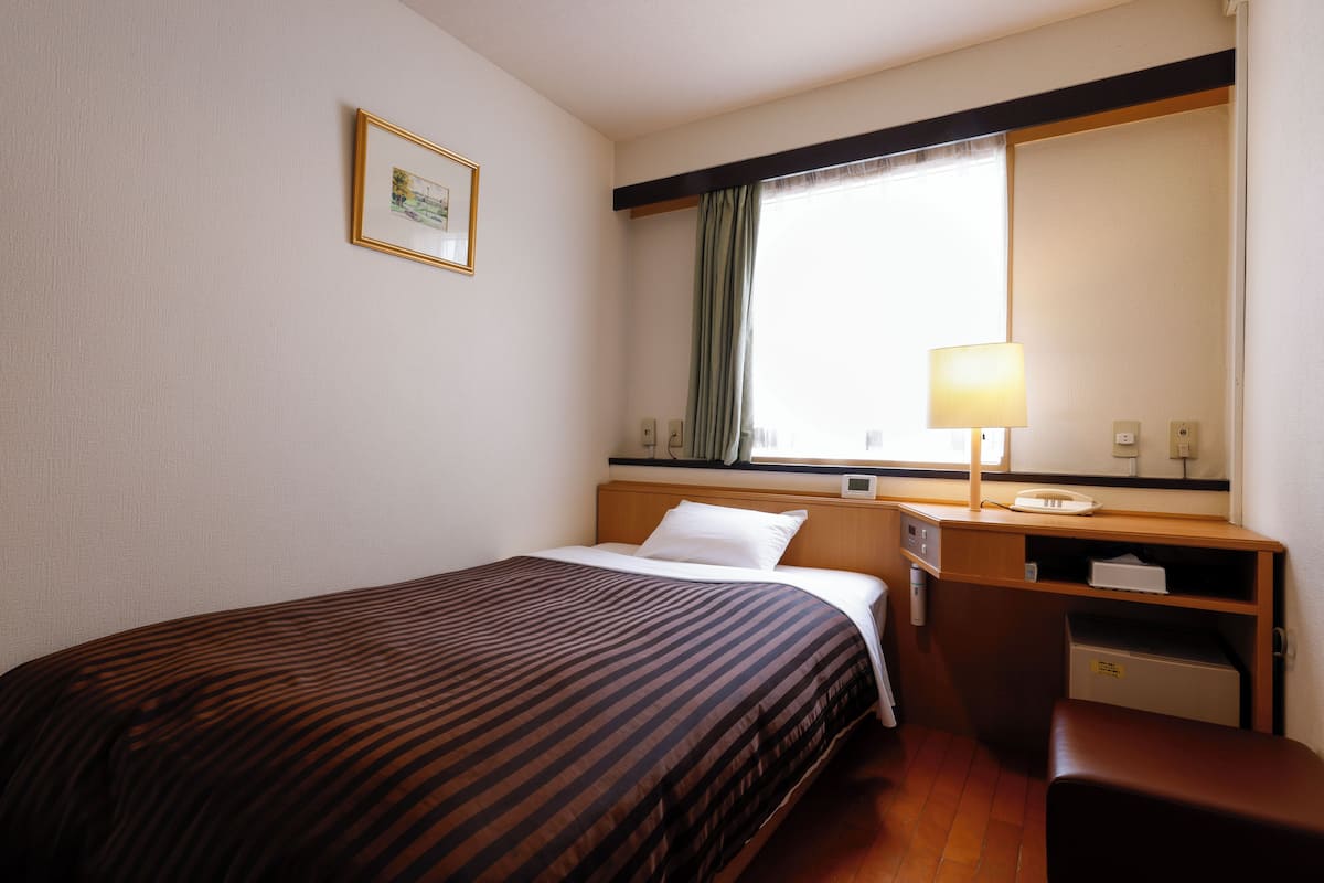A hotel room with a bed, a desk with a lamp, a chair, and a window with curtains.