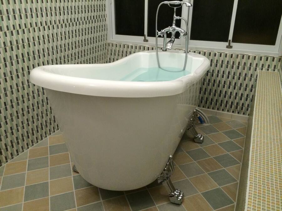 A white freestanding bathtub in a bathroom with patterned tiles and a window above it.