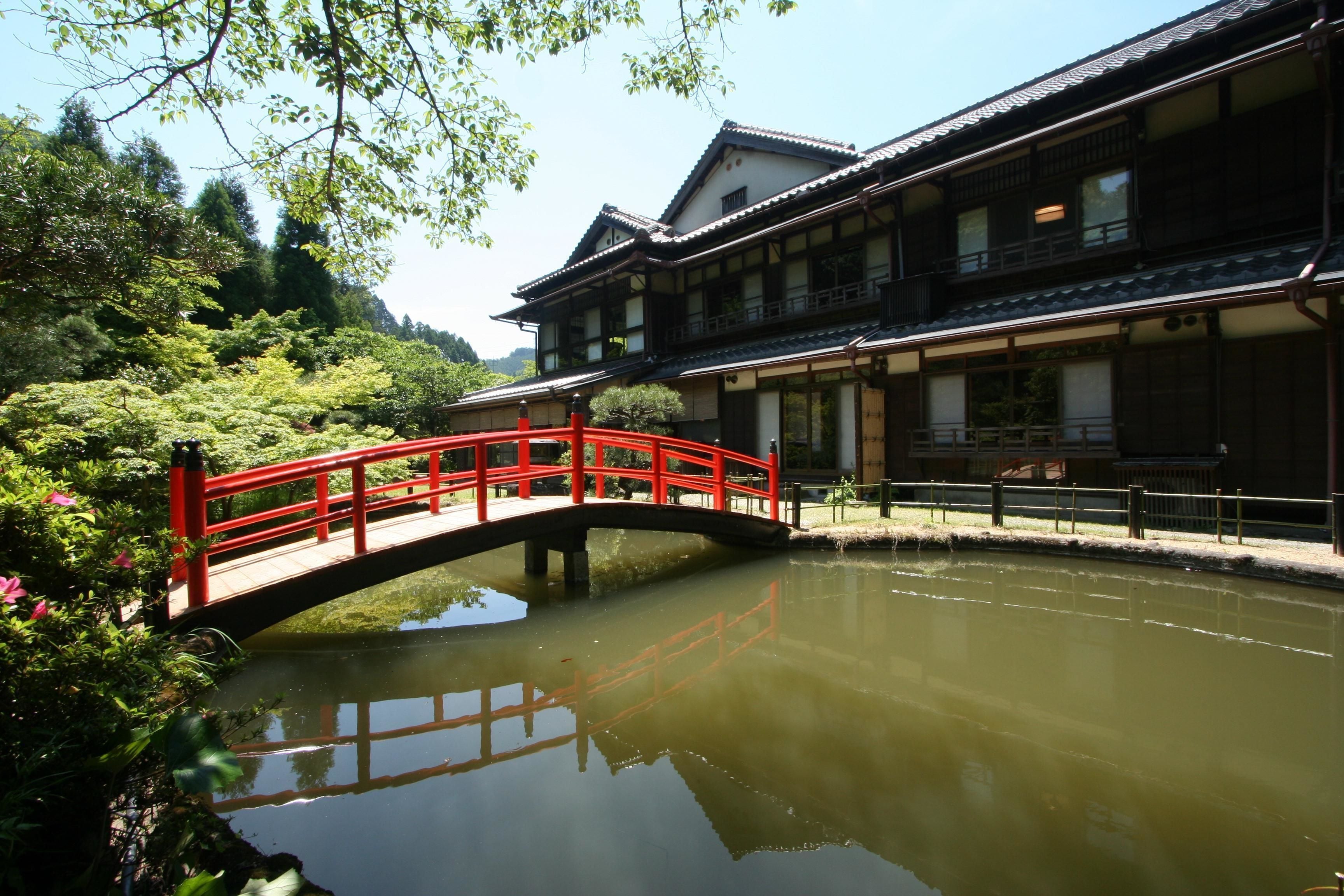 A traditional building with a red bridge over a pond, surrounded by greenery.