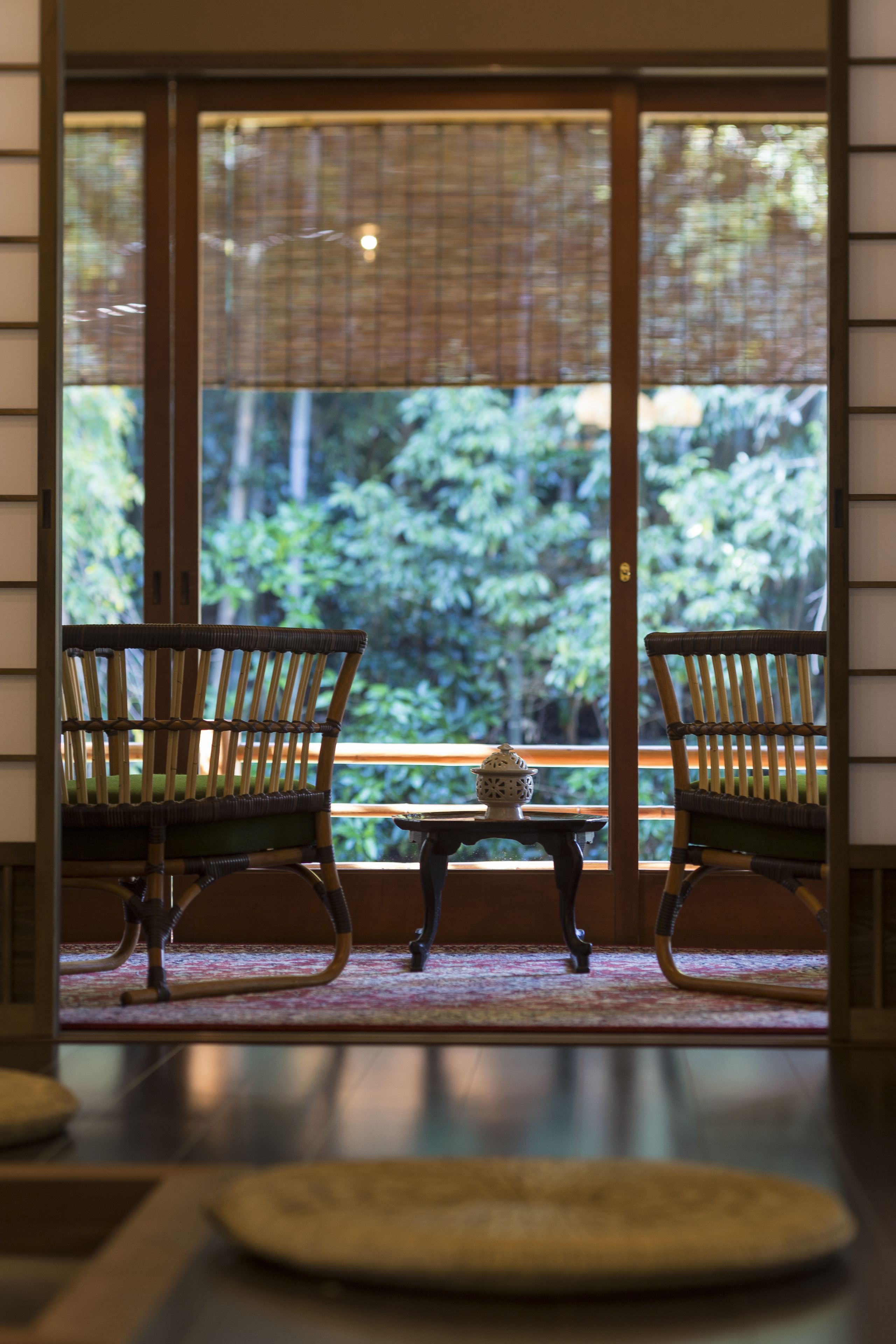 A room with two wicker chairs facing a large window, a small table with a decorative item, and a view of greenery outside.