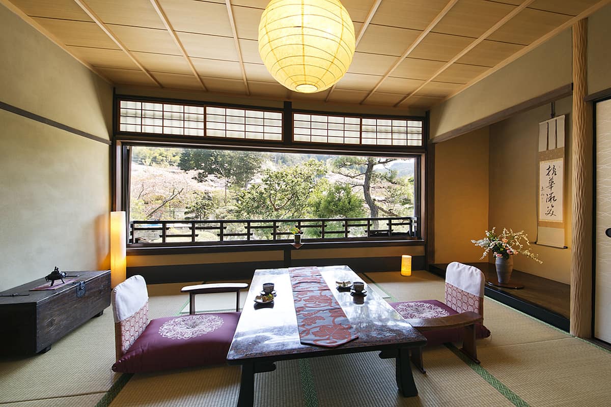 A traditional Japanese room with tatami flooring, a low table with a table runner, a cushioned bench, a hanging paper lantern, and a view of cherry blossoms through a shoji screen.